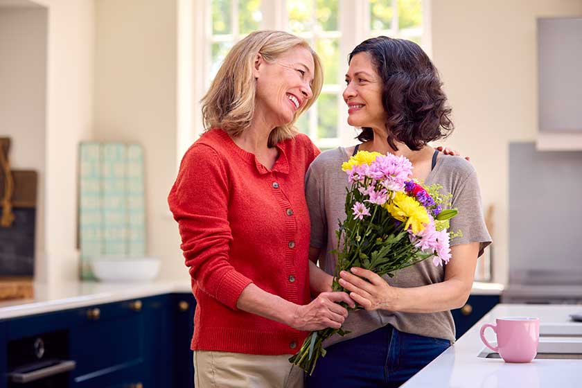 Loving Same Sex Mature Female Couple In Kitchen With Woman Giving Partner Flowers